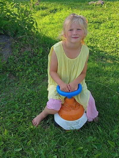 The image shows a young girl with blonde hair sitting on an orange and white ride-on toy in a grassy outdoor area. She is wearing a yellow pleated dress with pink translucent ruffles on the sleeves and legs. She is holding onto a blue handle of the toy. The girl is smiling at the camera, appearing happy and relaxed. The setting is a sunlit lawn, suggesting it is daytime with good weather. The grass is lush and green, with some weeds and small yellow flowers visible. The overall mood of the image is cheerful and captures a moment of childhood play. The location context indicates the scene is in Ostrovtsy, Russia. There is no visible text in the image.