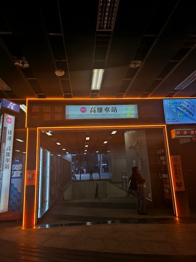 The image depicts the entrance to the Kaohsiung MRT (Mass Rapid Transit) Kaohsiung Main Station, specifically an exit. The architecture features a prominent neon orange illuminated frame around the entranceway. Above the entrance, a large illuminated sign displays "R11 高雄車站 2," indicating it's station R11, Kaohsiung Main Station, and possibly exit number 2. Below this, smaller text reads "Kaohsiung Main Station" and "Entrance." To the left, a vertical sign with blue and white lettering says "捷運 高雄車站," translating to "MRT Kaohsiung Station." To the right, another illuminated sign displays Japanese text and the logo "MOFAN," with smaller text in Chinese. A person wearing a backpack stands near the entrance, facing away from the camera and towards the interior of the station. The ground appears wet, reflecting the orange and blue lights. Stairs are visible leading down into the station. The overall environment is dimly lit, characteristic of an evening or night scene at a transportation hub in Kaohsiung, Taiwan.