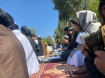 In the city of Arghandab, Afghanistan, a large group of men is observed outdoors, seated in rows on woven mats. Approximately two dozen individuals are clearly visible in the foreground and midground, with more suggested in the distance. The men are predominantly wearing traditional attire, including long robes (shalwar kameez) in colors such as brown, white, black, and grey, along with turbans and head coverings, mostly dark. Many individuals possess beards. They are seated on patterned mats, primarily reddish-brown, with their legs folded. Several individuals have their hands raised or clasped, consistent with an act of collective prayer or supplication; some appear to be holding prayer beads. The scene is illuminated by bright daylight under a clear blue sky. Sparse trees with green foliage are visible in the midground and background. Distant hills or terrain are faintly visible on the horizon. No direct interaction between individuals is discernable; the collective action appears synchronized. Many individuals are barefoot.