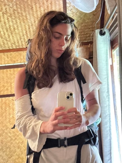A woman with long, wavy brown hair lies in a room with woven straw walls. She wears a white shirt and what appears to be a harness with black straps and buckles. She holds a light-colored smartphone in her hand, possibly taking a selfie. The background includes a partially visible fan, a window with light filtering through curtains, and a metal bar above the woman.