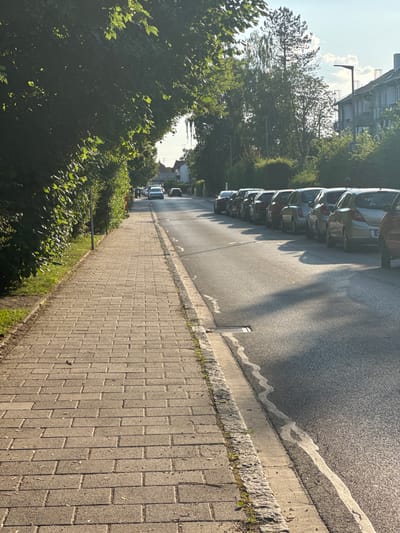 This image depicts a street scene in Höhenkirchen-Siegertsbrunn, Germany, likely during the daytime given the bright sunlight casting distinct shadows. The perspective is from a sidewalk looking down a street lined with parked cars and lush green trees.

The primary subjects are a row of parked cars on the right side of the street, appearing to be a mix of sedans and possibly an SUV. The cars are facing away from the viewer, suggesting they are parked parallel to the road. To the left of the street, a paved sidewalk runs parallel to the road, bordered by a curb and then a strip of grass and dense green foliage. Beyond the foliage, the branches of trees, some with visible leaves, create a canopy. In the background, a multi-story building with a distinct architectural style, possibly apartments or offices, can be seen peeking through the trees. Further down the street, more buildings and trees are visible, hinting at a residential or mixed-use area.

The activity is static; it's a view of a parked street. The time of day is suggested to be late afternoon or early evening by the long shadows stretching across the road and sidewalk. The weather appears to be clear and sunny. There is no visible text in the image. The overall mood is serene and ordinary, capturing a typical urban or suburban streetscape on a pleasant day.