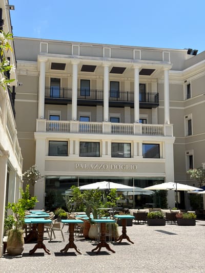 The image depicts the outdoor seating area of the Palazzo Dòglio in Cagliari, Italy. The facade of the building features neoclassical architecture with prominent white columns, balconies, and large windows. Below the windows, the name "PALAZZO DOGLIO" is clearly visible. The outdoor space is paved with cobblestones and furnished with numerous tables and chairs, some with turquoise tops and others with wicker seating. Several large white umbrellas provide shade over some of the seating arrangements. Green plants, including potted trees and cacti, are strategically placed around the seating area, adding a touch of nature to the scene. In the background, glimpses of indoor spaces are visible through the large windows, suggesting a restaurant or hotel setting. The sky is clear and blue, indicating a sunny day. There are no people visible in the image.