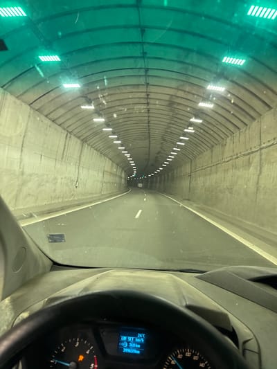 The image shows the interior of a vehicle driving through a tunnel in Golemo Buchino, Bulgaria. The tunnel has a curved ceiling with rows of lights. The walls of the tunnel are made of concrete. The dashboard of the vehicle is visible in the foreground, showing a digital display with the temperature as 24°C and the speed as 0 km/h. The odometer shows 399659 km. The road ahead is paved and has white lane markings. A white vehicle can be seen in the distance within the tunnel.
