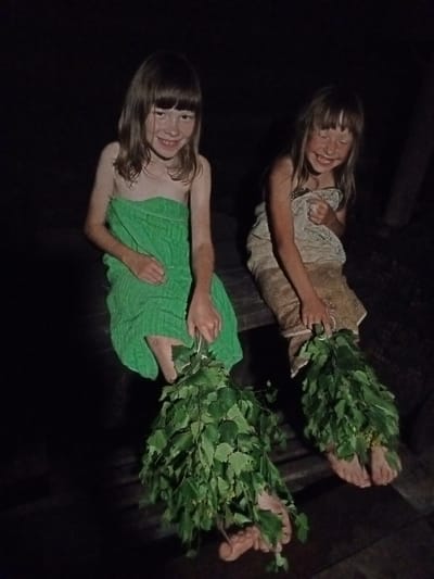 Two young girls are seated on a wooden bench. The girl on the left is wearing a green towel and holding a bundle of leafy branches in her right hand. The girl on the right is wearing a patterned cloth garment and holding a similar bundle of leafy branches in her left hand. Both girls are smiling. The background is dark and indistinct, suggesting an indoor or nighttime setting. There is a wooden structure beneath their feet, possibly part of a sauna or bathhouse. The overall atmosphere appears relaxed and joyful. The location of Buraly, Russia, is mentioned in the prompt as context for the image.