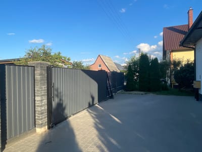 The image depicts an outdoor scene on a sunny day, likely in a residential area of Мухавецкі сельскі Савет, Belarus. In the foreground, a gray corrugated metal gate with brick pillars stands closed. The ground in front of the gate is paved with light gray interlocking bricks, with long shadows cast across it from the gate and unseen objects. To the right of the gate, a paved driveway leads towards a yellow house with a brown tiled roof and a red brick chimney. Behind the gate, there are trees and smaller buildings, including a distinctive red house with a grey tiled roof and a single small window. The sky is a clear blue with a few white clouds. Power lines are visible stretching across the upper portion of the image. The scene appears to be a private property entrance.