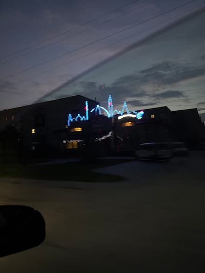 The image captures a nighttime view of a building in Berezhany, Ukraine, adorned with illuminated decorations. The building appears to be a multi-story structure with several windows, some of which are lit from within. The most prominent feature is the neon-like blue and red lights outlining what seems to be a mosque or a similar architectural style, complete with minarets and domes, running across the facade of the building. This decorative lighting creates a striking contrast against the dark evening sky and the silhouetted building.

The sky itself is a deep twilight blue with some clouds visible. Overhead, power lines are strung across the frame, a common sight in urban environments. In the foreground, the blurred silhouette of a car's side mirror and part of its hood suggest the photograph was taken from inside a moving vehicle. The road surface is visible in the lower part of the image, also showing motion blur. Two vehicles, one appearing to be a van or minivan, are parked or moving slowly on the street in front of the building. The overall impression is of a street scene at dusk or early night, with the building's festive lights drawing the eye.