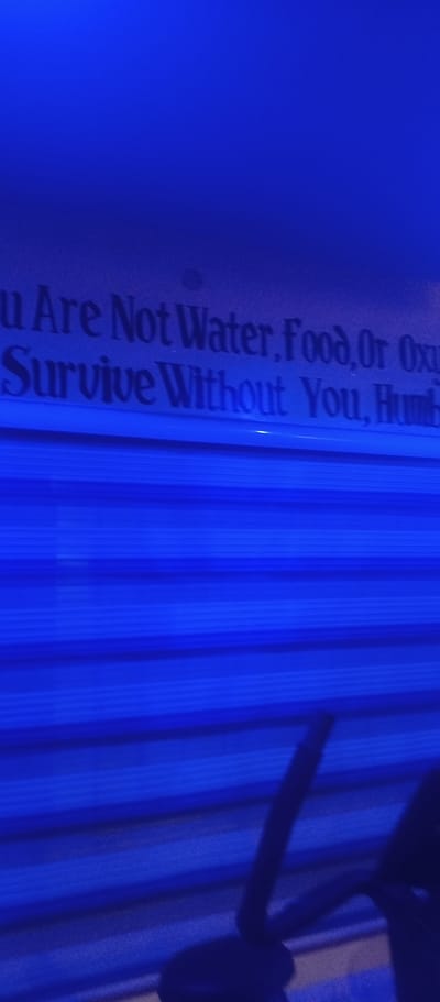The image shows text on a blue background. The text reads: "u Are Not Water, Food, Or Oxygen, We Cannot Survive Without You, Humbling." The text appears to be a sticker or decal. The background is a ribbed blue surface, possibly part of a tanning bed or a similar structure. There are no people visible in the image. The location in Karu, Nigeria is not discernible from the image.