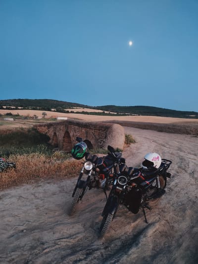 Two motorcycles are parked on a dirt path in front of an old stone bridge. The motorcycles are black with red and white accents. One motorcycle has a green and black helmet on it, and the other has a white helmet with pink accents. The bridge has three arched openings and is made of stone. Behind the bridge, there are dry, golden fields and a line of trees on a hill. The sky is a deep blue with a crescent moon visible. The scene suggests it is dusk. The location is Filipeni, Moldova.