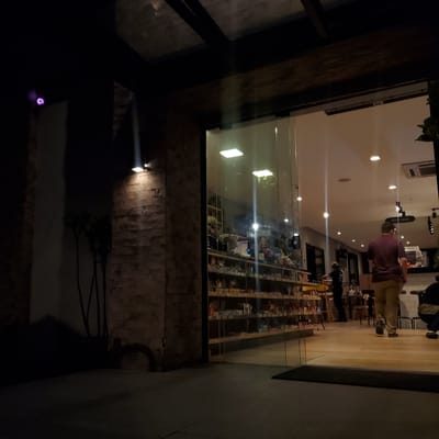 Here is a description of the image:

The image captures a nighttime scene, seemingly taken from an outdoor perspective through a glass window or door, into a cafe or shop in Curitiba, Brazil. The interior is brightly lit, showing customers browsing shelves filled with items. One person is facing away from the camera. The outside area is dark, with visible brickwork and what appears to be a dark object resembling a tire against the wall, along with some wiring. Two lights on the exterior illuminate the brick wall.
