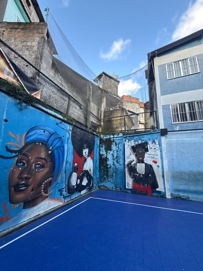 This image captures an outdoor community sports court within a favela in Rio de Janeiro, Brazil. The foreground features a bright blue modular sports court surface with white and red demarcation lines. Surrounding the court are walls painted blue, adorned with three distinct murals.
On the left wall, a large mural depicts a Black woman from the chest up, with dark skin, a blue headwrap, large pearl hoop earrings, and intricate blue and white makeup. Orange leaf-like designs frame her head. Adjacent to this, a second mural portrays a person with an afro hairstyle, shirtless, wearing boxing gloves, set against a red background. The wall section between the murals and on the right shows wear, including peeling paint and some holes. On the right wall, a third mural features a young Black individual in a graduation cap and gown, holding a microphone. The background of this mural includes stylized architectural drawings and Portuguese text, notably "O que você quer ser quando crescer?" (What do you want to be when you grow up?).
Above the walls, a dark mesh safety net spans the area, held by poles and connected to surrounding structures. In the background, a dense urban landscape of multi-story buildings is visible under a clear blue sky with scattered white clouds. Buildings vary in color, including light green, grey, brick red, and shades of blue. The building on the far right is blue and light blue, with multiple windows fitted with white security bars. Various architectural elements like ledges, satellite dishes, and exposed brickwork are present on the background structures.