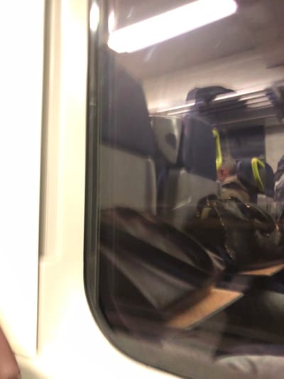 The interior of a train car is visible through a slightly grimy window. Seating includes rows of blue-gray seats, with luggage or personal items stored in overhead compartments. A person wearing a yellow and black hat is seated. A black backpack sits nearby. The window frame is black and the interior wall is white. Bright artificial lighting illuminates the scene. The image appears to be taken from inside another train car.