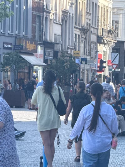 This is a medium shot of a street scene in Bruxelles, Belgium, taken during the day. The image is oriented vertically, showing a bustling street lined with multi-story buildings. The perspective is from a slightly elevated viewpoint, looking down the street.
Several people are visible, some walking and others standing. In the foreground, a woman wearing a light yellow dress and carrying a black bag on her back is partially visible. To her left, another person is seen from the back, wearing a white shirt and blue jeans. Further back, more individuals are dispersed along the street, suggesting a lively atmosphere.
The buildings lining the street exhibit a mix of architectural styles, with some appearing older and ornate, featuring balconies and detailed facades, while others are more modern. Various signs are attached to the buildings, indicating businesses or services. Visible text includes "ATM", "DRINKS CORNER", and "Belgastra". The street is paved with cobblestones, and trees are present, adding greenery to the urban environment. The time of day appears to be daytime, with bright lighting casting shadows, suggesting it could be late morning or afternoon. The overall mood is one of everyday city life.