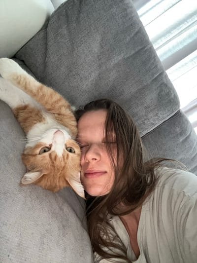 The image shows a person and a cat lying on a grey sofa. The person is lying on their back, with their head resting on the sofa. The cat is lying next to the person, with its head resting on the person's chest. The cat is orange and white, and it is looking up at the person with its eyes open. The person is wearing a light-colored t-shirt. The background is a grey sofa with some light coming from the right side, possibly from a window. The overall scene suggests a moment of relaxation and companionship. This scene is not specific to Baan Hua Thanon, Thailand, as it depicts a common domestic setting.