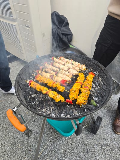 A grill filled with skewers is located outdoors. The skewers contain meat and vegetables, some with a yellow coating. The grill is round and black. The ground is gray with small pebbles. A blue cart sits next to the grill, and grilling tongs are positioned near the cart. A white air conditioning unit is adjacent to the grill. People are standing around the grill, visible by their clothing and shoes. The location is Tourcoing, France.