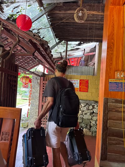 Here is a description of the image:
The image captures a person standing within a building in what is described as Mèo Vạc Commune, Vietnam. The individual, who is wearing a gray shirt and light-colored shorts, has a black backpack and is holding onto two black luggage cases with a blue trim.
The interior of the building features a mix of construction materials, including wood, stone, and brick. Decorations such as a red lantern and woven lamps are visible. Wall-mounted signs with Vietnamese text are present. The building's structure is partially open to the outside, showcasing foliage and other buildings in the background.