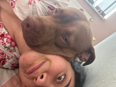 This is a close-up, selfie-style photograph taken indoors, likely in a bedroom, given the presence of a bed with floral-patterned bedding and a hint of what appears to be a radiator and blinds in the background. The primary subjects are a woman and a brown dog, possibly a pit bull mix, nestled closely together. The woman's face is partially visible, with her eyes wide and looking upwards, and her lips pursed in a playful or surprised expression. Her dark hair is also visible. The dog's head is pressed against the woman's face, with its ear visible and its brown fur prominent. The lighting suggests it is daytime, possibly morning or afternoon. The overall mood is intimate and affectionate, capturing a moment of connection between the woman and her pet. There is no visible text in the image. The location context provided is Malevo, Bulgaria.