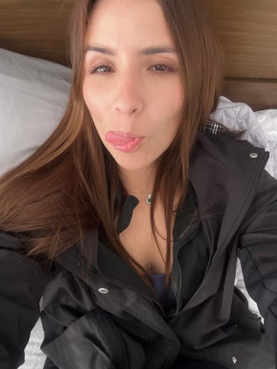 A young woman lies on a gray pillow. She sticks out her tongue slightly. Her hair is medium brown, and she has freckles on her nose. She wears a black puffy jacket. A silver necklace with a green pendant rests on her chest. The image is taken from an overhead perspective with the camera rotated.
