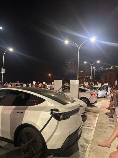 The image depicts a night scene at a Tesla charging station in Alicante, Spain, with fireworks visible in the background. The foreground is dominated by a white Tesla Model Y, plugged into a charging station. Its rear taillights are illuminated red. Other Teslas and a silver sedan are also present, parked in charging spots. A crowd of people, dressed casually, are gathered around the charging stations, some looking towards the fireworks. The environment appears to be an outdoor public space with streetlights and buildings in the distance, illuminated by the fireworks. The overall atmosphere suggests a festive occasion.
