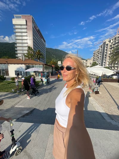 In the city of Gagra, Abkhazia, a blonde woman with sunglasses is taking a selfie in a coastal setting. She is wearing a white tank top and beige shorts. Behind her, a promenade is lined with shops and umbrellas. In the background, there are modern buildings, palm trees, and lush green mountains under a bright blue sky with scattered clouds. People are seen walking along the promenade, and a child is on a scooter in the foreground. The overall scene depicts a sunny day at a resort town.