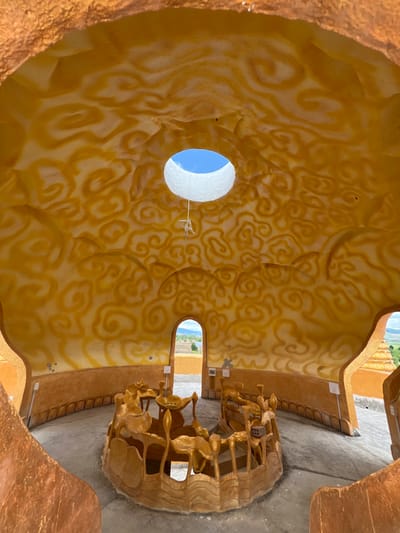 The interior of a circular, domed structure in Đăk Mar, Vietnam, is captured from a low-angle perspective. The ceiling and upper walls are a textured, molded surface painted in an earthy orange-brown base with a lighter yellow-orange swirling, cloud-like pattern. A circular oculus at the dome's apex reveals a clear blue sky, framed by a prominent white cylindrical lip. A white, knotted string hangs from the center of the oculus. The lower walls are a darker brown, featuring a simple base molding. The floor is a flat, light gray concrete surface. Centrally located on the floor is a large, circular, sculpted structure painted in a uniform golden-brown. This central piece features multiple organic, stylized elements resembling lotus flowers, leaves, and tiered basins, configured as a dry fountain or altar. Two arched doorways are visible in the far wall, opening to a brightly lit exterior landscape of green vegetation and distant hills under a blue sky. No individuals, actions, or interactions are present within the scene.