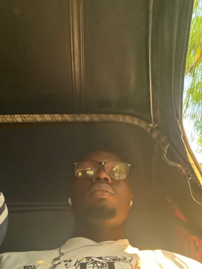 The image features a close-up of a young person wearing glasses and a white hoodie with a black design. The person is lying down, with their head tilted upwards and their eyes closed or looking away. Sunlight is casting a warm glow on their face and shoulder, suggesting it is daytime. The background appears to be the interior of a vehicle, possibly a car, with dark upholstery and a glimpse of greenery reflected on the ceiling, indicating an outdoor scene. The person's relaxed posture and the soft lighting create a serene and peaceful atmosphere. The overall impression is one of a moment of rest or quiet reflection, possibly during a car ride. The location context provided, Amansee, Nigeria, suggests the setting is within this region. No visible text is present in the image.