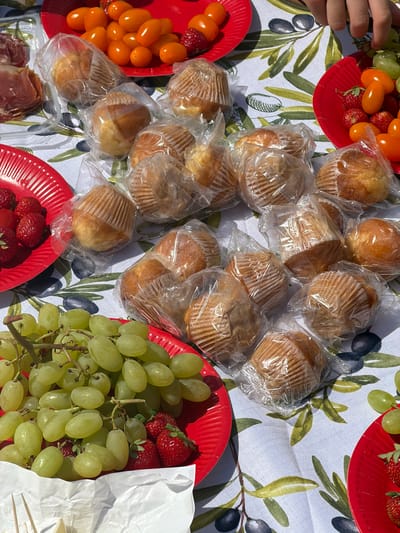 This image depicts a picnic spread on a patterned tablecloth, likely outdoors due to the bright sunlight casting shadows. The scene is dominated by a variety of food items arranged on red paper plates and a clear plastic container. Visible foods include numerous individually wrapped muffins, a generous bunch of green grapes, bright red strawberries, and small, orange cherry tomatoes. There's also a portion of thinly sliced cured meat, possibly ham or prosciutto, in the plastic container. A hand, belonging to an unseen person, is visible reaching towards the upper left portion of the frame, suggesting active participation in the picnic. The tablecloth features a repeating pattern of olive branches with green and dark blue olives. The overall impression is one of a casual, enjoyable gathering, likely a picnic or an outdoor meal. The lighting suggests it is daytime, and the presence of fresh food and a relaxed arrangement implies a pleasant, leisurely occasion. The location is specified as Trouville-sur-Mer, France, which suggests a possible coastal setting, though not explicitly visible in the image itself. No visible text is present in the image.