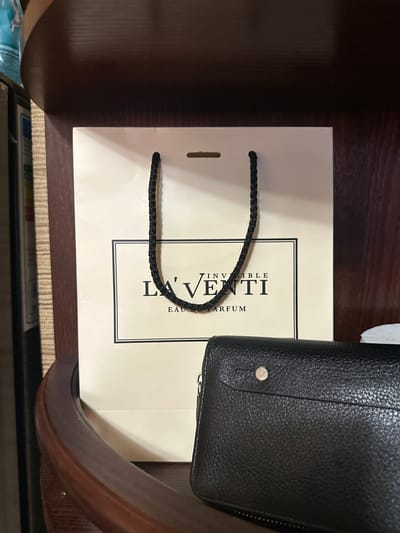 A light-colored shopping bag with black braided handles sits on a wooden shelf. The bag features a black-bordered rectangular logo with text that reads "LA'VENTI INVISIBLE EAU DE PARFUM". In front of the bag, a black textured wallet with a gold button closure is visible. The background is a dark wooden surface. The image appears to be taken indoors, possibly in a home or retail setting in Aleksandrovka, Russia.