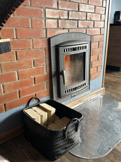 In Listvyanka, Russia, a basket of compressed sawdust fire logs sits on a metallic hearth in front of a brick fireplace. The fireplace has a cast iron insert with a glass door, through which a small flame is visible. The brickwork is rustic red with visible mortar. A black brush with dark bristles is positioned to the left of the fireplace, partially visible. The flooring is dark wood planks. The overall scene suggests a cozy, indoor setting, likely a home or guesthouse, designed for warmth and comfort.