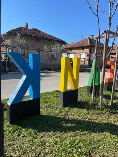 The image shows a street scene in Dolni Vit, Bulgaria. Large, colorful, block letters - blue "K", yellow "N", green "A", orange "N", are situated on the grass next to a sidewalk. Buildings are visible in the background with tiled roofs and brick or stucco walls. A network of tree branches is in the top left corner. The sky is blue and clear.