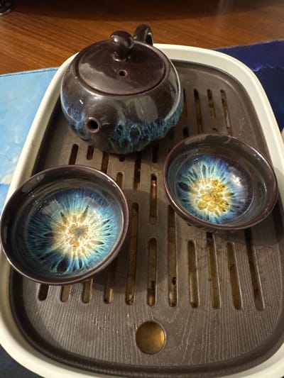 The image shows a tea set consisting of a teapot and two cups placed on a tea tray. The teapot is dark brown with blue and gold glazes, and it has a round lid with a small knob. The cups are also dark brown with a similar glaze pattern on the inside, resembling a celestial explosion or a flower. The tea tray is dark gray with horizontal slits and a circular indentation on the right side. The setting appears to be indoors, with a wooden surface visible on the left and a blue fabric beneath the tea tray. The lighting is warm, suggesting artificial light. There is no visible text in the image. The overall impression is of a serene and aesthetically pleasing tea ceremony setup, possibly in a home environment in Shcherbinka, Russia.