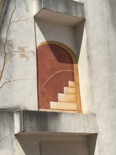 The image is a close-up of a mural painted on the exterior wall of a building in Mercurol-Veaunes, France. The mural depicts a stylized scene with a tree on the left side, rendered in brown and grey tones against a textured, off-white background. To the right of the tree, an arched entryway is painted, with a dark reddish-brown interior. Inside the entryway, a set of cream-colored stairs with yellow detailing ascends, and a cream-colored handrail curves along the side. The architectural elements of the building, such as concrete ledges and the textured surface of the wall, are also visible and are showing signs of age with water stains and some cracking. The lighting suggests it is daytime, with shadows cast by the architectural features and the painted elements. There are no people visible in the image.