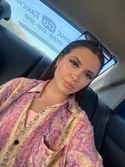 A young woman is seen from the chest up, taking a selfie inside a vehicle. She is wearing a long-sleeved, collared shirt with a snakeskin-like pattern in shades of pink, purple, and yellow. She has on a peach-colored tank top underneath. Her hair is dark brown and pulled back, with sunglasses perched on top of her head. The background shows the interior of a car, with a black leather seat visible on the right side and the rear window of the car on the left. The rear window has text on it, which is partially obscured, but appears to be an advertisement or logo. The scene is lit by natural light. There is no indication of the location being in Gelinbatan, Russia.
