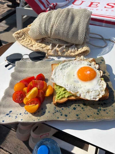 The image depicts a close-up, overhead view of a breakfast spread on a white table, likely at an outdoor venue in Lozenets, Bulgaria. The meal consists of a slice of toast topped with mashed avocado and a fried egg, accompanied by a side of halved cherry tomatoes (both red and yellow varieties). The food is served on a speckled ceramic plate. To the left of the plate are a pair of silver-rimmed sunglasses with dark lenses, resting on a straw-like woven mat. Behind the mat, a light-colored mesh bag with fringed edges is partially visible. In the background, a red and white striped towel or blanket is draped over a white chair. Sunlight casts distinct shadows on the table and the wooden surface beneath. The overall scene suggests a leisurely breakfast, possibly by the sea given the light and relaxed setting.