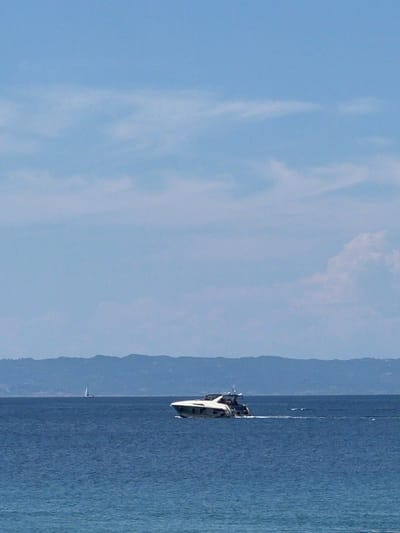 A bright, daytime, medium-shot captures a wide expanse of the Aegean Sea, with a distant, hilly coastline forming the horizon under a pale blue sky dotted with sparse clouds. In the foreground, the deep blue water shows gentle ripples. A white motor yacht with a dark hull is the primary subject, seen from its port side as it moves from left to right, leaving a faint wake. Further to the left and smaller in scale, a solitary white sailboat is visible. The scene appears to be set in the waters off the Sithonia Municipal Unit in Greece, characterized by its clear waters and characteristic coastal topography. There are no people clearly visible on either vessel.