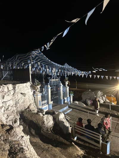 A nighttime scene features a building facade on a rocky cliffside. The structure is decorated with blue and white striped fabric and triangular flags. A person sits on a white bench on a small balcony, overlooking the dark surroundings. A drum sits on a wall. The light overhead highlights the decorations and the textures of the rock face.