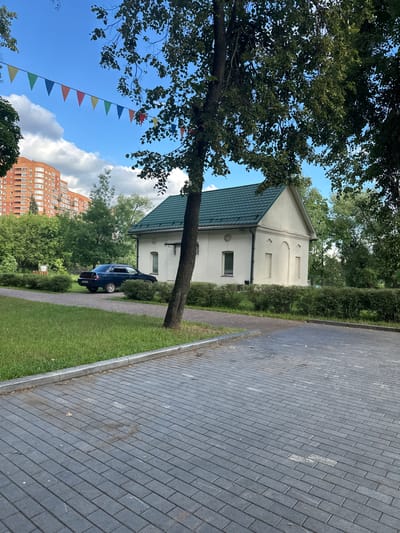 In Kotelniki, Russia, a dark blue sedan is parked on a paved driveway next to a small, white building with a green roof. The building has two arched niches on its facade and a single window on the left. A mature tree with dark green leaves stands in front of the building. The driveway is constructed of gray interlocking pavers, bordered by a strip of green grass and a concrete curb. In the background, a tall, orange residential building with many windows is visible above a line of lush green trees. A string of colorful triangular flags hangs across the sky. The sky is blue with scattered white clouds, and the overall scene appears to be a sunny day in a residential area.