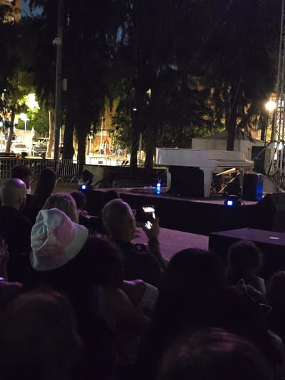 This media file depicts an outdoor event at night, likely a concert or performance, with a piano and audience members visible. The setting appears to be a public space, possibly a park or plaza, indicated by the presence of trees, lighting structures, and fencing.

The main subjects are the people attending the event. They are seen in various positions, some looking towards the stage area, others engaged with their phones. The crowd is dense in some areas, suggesting a popular event.

In the background, a white grand piano is positioned on a stage. A performer, partially obscured, appears to be playing the piano. Lighting elements, including blue lights on speakers and stage lights, contribute to the nighttime ambiance. The scene is dimly lit, with spotlights highlighting the performance area and casting shadows.

Notable details include the time of day, which is clearly night due to the darkness and artificial lighting. The presence of trees suggests an outdoor location. The overall emotion conveyed is one of engagement and enjoyment, with people gathered to watch and listen to the performance.

There is no visible text in the image. The context provided indicates the location as Cornellà de Llobregat, Spain.