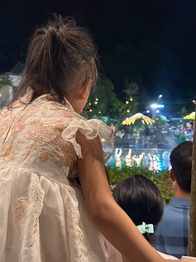 An over-the-shoulder shot captures a young girl, seen from the back and right side, looking towards a nighttime outdoor event. She is seated on someone's shoulders, with her dark, messy hair tied up in a bun. She is wearing a light-colored, embroidered dress with floral patterns in pink and gold. The dress has ruffled sleeves. To the right of the girl, another child's head with dark hair and a light blue hair clip is visible. Further to the right, a man in a blue plaid shirt is also facing the event.
The background is dark, indicating nighttime, with blurred lights and trees. In the distance, a performance or display is taking place, with several figures in bright, colorful costumes visible in a shallow body of water, possibly a canal or pond. Above them, illuminated decorations, likely strings of lights, are visible, creating a festive atmosphere. The scene appears to be in a public outdoor space, possibly a park or festival area in Hoa Lư Ward, Vietnam, given the context. The overall impression is of a family enjoying an evening event.