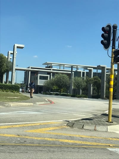 The image displays an urban scene in Sandton, South Africa, under a clear blue sky with sparse clouds, indicating bright daylight. In the foreground, an asphalt road features yellow double solid lines, yellow zigzag lines, and white dashed lane markings. A concrete curb, partially painted red and white, delineates the road edge. To the right, a yellow pole with black horizontal stripes supports a traffic light showing a red illumination.
In the mid-ground, an adult male walks on a paved pathway, moving away from the viewer. He is dressed in a dark blue jacket, light brown trousers, and dark shoes. To the left of the pathway, a grassy slope is bordered by a trimmed green hedge.
The background is occupied by modern, multi-story buildings constructed primarily from concrete and glass. These structures exhibit a distinctive architectural style characterized by prominent vertical pillars and large, flat, cantilevered rooflines or overhead structures. Some building sections include horizontal slatted facades. The area around the buildings is landscaped with numerous small, round-canopy trees and green shrubbery. A light-colored vehicle is partially visible in the distance, behind the pedestrian, near what appears to be a building entrance. No direct interactions are discernible.