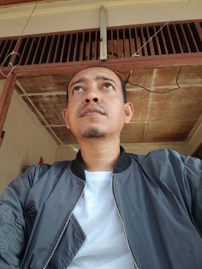 A person looks upward. They wear a white t-shirt and a gray bomber jacket. The background is a ceiling with brown-painted wooden rafters and a ventilation panel. Electrical cords are also visible. The lighting is even.