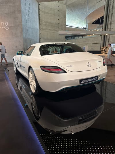 This image captures a white Mercedes-Benz SLS AMG Electric Drive parked on a mirrored surface, possibly within a museum or showroom in Stuttgart, Germany. The car is the primary object, its sleek design highlighted by the reflective floor which also mirrors a second, darker car. The setting appears to be indoors, with a concrete wall visible in the background, featuring embossed numbers and text. A portion of the wall also displays a vertical arrangement of small, circular lights.
Two people are present, both men, seen from the back. One is closer to the foreground, wearing a white t-shirt and shorts, looking down. Another figure is further back, near some machinery. The lighting suggests it's daytime, though the exact time is not discernible. The overall mood is calm and observational, with the focus on the automotive display.
Visible text includes "SLS" and "AMG" on the car's rear. The phrase "Electric Drive" is also visible on the car's side. The embossed numbers on the wall are not clearly legible.