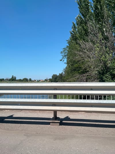 The image shows a guardrail along the side of a road, with a body of water and a distant treeline visible beyond it. The sky is a clear blue. The guardrail is made of silver metal and has three horizontal bars. The road surface is visible in the foreground, and shadows are cast by the guardrail and its supports. To the right, a large tree with green leaves and some bare branches is prominent. The background shows a row of trees, houses, and a small body of water. The overall scene suggests a sunny day, likely in a rural or suburban area. The location is identified as Ильинка, Russia.