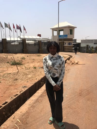 The image depicts an outdoor scene in Mista Ali, Nigeria, specifically at the entrance to AG Medical Services. A young female stands centered in the foreground, facing the camera with a neutral expression. She has dark, braided hair and wears a long-sleeved, collared shirt with a grey and white camouflage pattern, dark trousers, and light green slides. Her hands are clasped in front of her waist.
The ground is unpaved, consisting of reddish-brown dirt and sparse dry vegetation. A rectangular drainage ditch constructed from concrete blocks runs along the left side of the unpaved road. In the midground, a low concrete fence topped with what appears to be barbed or electric wire extends across the scene. Behind this fence, several light-colored buildings with metal roofs are visible. To the left, a line of at least seven flagpoles displays various flags, including the Nigerian flag.
To the right of the primary subject, a two-story gatehouse with a light-colored pyramidal roof serves as the entrance to the facility. A blue sign on the gatehouse reads "WELCOME TO AG MEDICAL SERVICES." A tall streetlight pole stands behind the gatehouse. Further along the unpaved road to the right of the gatehouse, a distant figure dressed in dark clothing walks away from the camera. The sky is clear and bright blue.