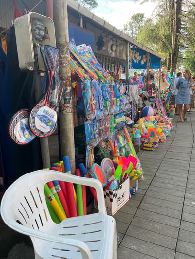 A street market stall in Ureki, Georgia, displays a variety of beach and water toys. The stall is set up outdoors on a wooden boardwalk, with a covered structure in the background that houses more merchandise. In the foreground, a white plastic chair is filled with colorful foam pool noodles in red, yellow, and green. To the right of the chair, a cardboard box overflows with brightly colored plastic shovels and rakes, along with other toys like a small yellow toy construction vehicle. Further down the stall, an assortment of water guns, goggles, and snorkels are neatly displayed on racks. Beach balls, including one large blue and white striped ball, are also visible. A woman in a denim dress is seen walking away from the camera, and another woman with blonde hair is visible in the background, suggesting a public area with pedestrian traffic. The overall impression is of a lively summer market catering to vacationers.