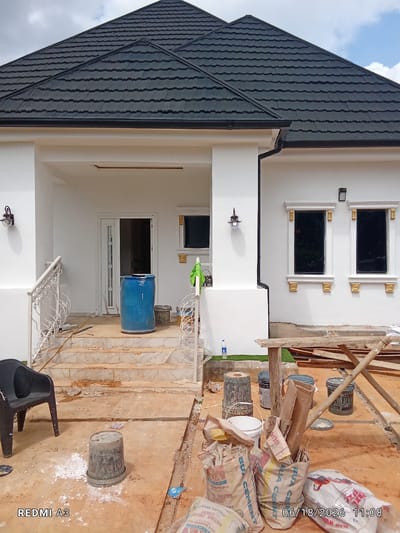 The image depicts the exterior of a white, modern house under construction in Nawfia, Nigeria. The house features a dark, textured roof with visible shingles. The entrance area has a covered porch with white decorative railings and a set of stairs leading up to a white door. A large blue barrel stands on the stairs. Two dark-tinted windows with ornate gold trim are visible on the front of the house.
The foreground of the image shows a construction site with an unpaved, reddish-brown ground. Various construction materials are scattered around, including several bags of cement, buckets, wooden planks, and a black plastic chair. A metal railing with decorative scrollwork lines the stairs. A bottle of water and some green artificial turf are visible to the left of the house. The overall scene suggests ongoing construction work, with the house appearing to be in its finishing stages.