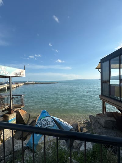 The image captures a section of the Balchik waterfront in Bulgaria, likely a marina or a beach area adjacent to a restaurant. In the foreground, a black metal railing separates the viewer from a rocky shoreline where a blue object, possibly a damaged inflatable or canvas cover, lies on the rocks. Graffiti is visible on this blue object, including the words "WATER PARADISE."
To the left, a portion of a structure with tables and chairs suggests an outdoor seating area for a restaurant or cafe. Above this area, a sign is partially visible, with the words "JET SAFARI" prominently displayed, indicating a recreational activity offered at this location. Further into the image, a pier extends into the turquoise water, lined with docked sailboats.
On the right side, a modern-looking building with large glass windows stands on stilts over the water, offering a clear view of the sea. The sky is a bright blue with scattered white clouds. The horizon shows a distant landmass, likely hills or mountains. The overall scene depicts a sunny day at a seaside location, with elements of leisure, recreation, and dining.