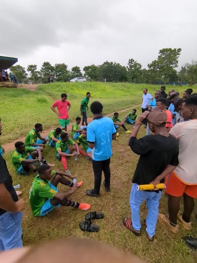 The scene takes place in a grassy field in Morogoro, Tanzania (latitude: -6.799, longitude: 37.63). A group of young men, likely a soccer team, is gathered on the field, some sitting and others standing. Many are dressed in green and blue soccer uniforms, with some wearing different clothing. A man in a white shirt and black pants is standing to the right of the group, seemingly addressing the players. A man wearing a red shirt and black shorts is standing to the left of the group, possibly a coach or team official. Several other men are standing around, some watching the players, others conversing. A yellow PVC pipe is lying on the ground near the feet of the man in the blue pants. The ground is uneven, covered in grass with patches of dirt. The sky is cloudy and overcast. The environment suggests an informal soccer training session or practice.