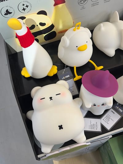 This image displays a collection of whimsical night lights, likely in a retail setting, possibly a store in Pekalongan, Indonesia. The subjects are primarily soft, silicone night lights shaped like cartoon animals. There's a white duck-like figure with yellow feet and beak, wearing a red knitted band around its neck. Another night light resembles a white bear with a black cross for an eye and pink cheeks. A third, partially visible, is a white figure wearing a purple hat. The background includes display packaging with text such as "informa," "Portable Night L," "INDOOR LIGHTING," and prices in Indonesian Rupiah (Rp). Also visible are phrases like "collectible series," "all night companion," "adjustable brightness," and "USB included," indicating product features. The overall scene suggests a display of children's or novelty lighting products, with a focus on cute and playful designs. The lighting is bright, suggesting an indoor environment, likely during daytime.