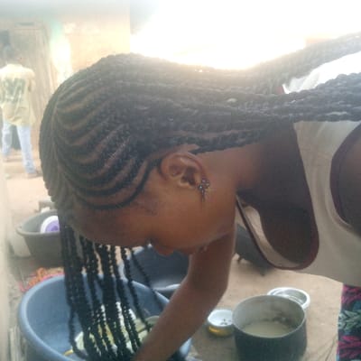 The image depicts an individual engaged in an activity outdoors in Ibreda, Nigeria. The primary subject, a person with dark skin, is presented in a left profile, bending forward over a dark blue plastic basin. The subject's hair is styled in tight cornrows on the scalp, which transition into numerous long, black box braids. The visible left earlobe features an intricate blue and purple beaded earring. The individual wears a light-colored sleeveless top with a dark trim and a patterned garment visible at the lower right. The subject's right arm is partially submerged into the blue basin, which contains a light-colored substance and green leafy items. To the right of the basin, a dark-colored pot holds a white, liquid substance, and a small white bowl contains dark granular material. In the mid-ground, a black plastic basin is partially visible. The background reveals a blurry figure of another individual in a light-colored shirt and blue jeans, standing near a light-colored building wall or doorway. The ground is composed of reddish-brown earth. The scene is illuminated by bright natural light originating from the upper left.