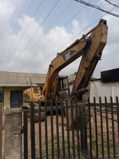 A yellow Caterpillar (CAT) 320C excavator is stationary on a dirt plot in Ijaiye, Nigeria, positioned behind a dark metal picket fence. The excavator's boom is raised, and its bucket rests near the ground. The machine exhibits signs of use, including dirt and wear on its surfaces. To the left of the excavator, a single-story building with a cream-colored facade and a grey corrugated metal roof features two dark blue doors. To the right, a structure partially covered in white sheeting is visible, with a darker structure behind it. The immediate foreground shows a dark metal fence with vertical slats, anchored by a concrete post on the left. The ground is primarily dirt with sparse green vegetation. Overhead, multiple parallel utility lines traverse a partly cloudy sky. No individuals are present, and no actions or interactions are depicted.